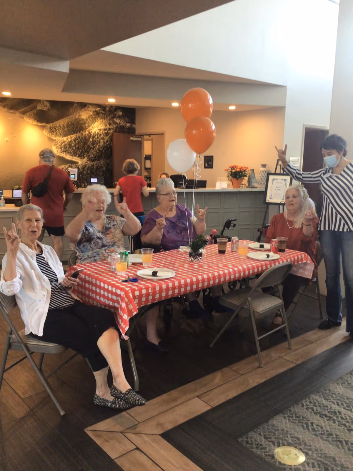 A group of elderly women sitting around a table with a red and white checkered tablecloth, enjoying drinks and snacks. Three balloons, two orange and one white, are tied to a small centerpiece on the table. In the background, a reception desk is visible with staff and other people. One woman standing to the right is wearing a mask and making a hand gesture along with the seated women.