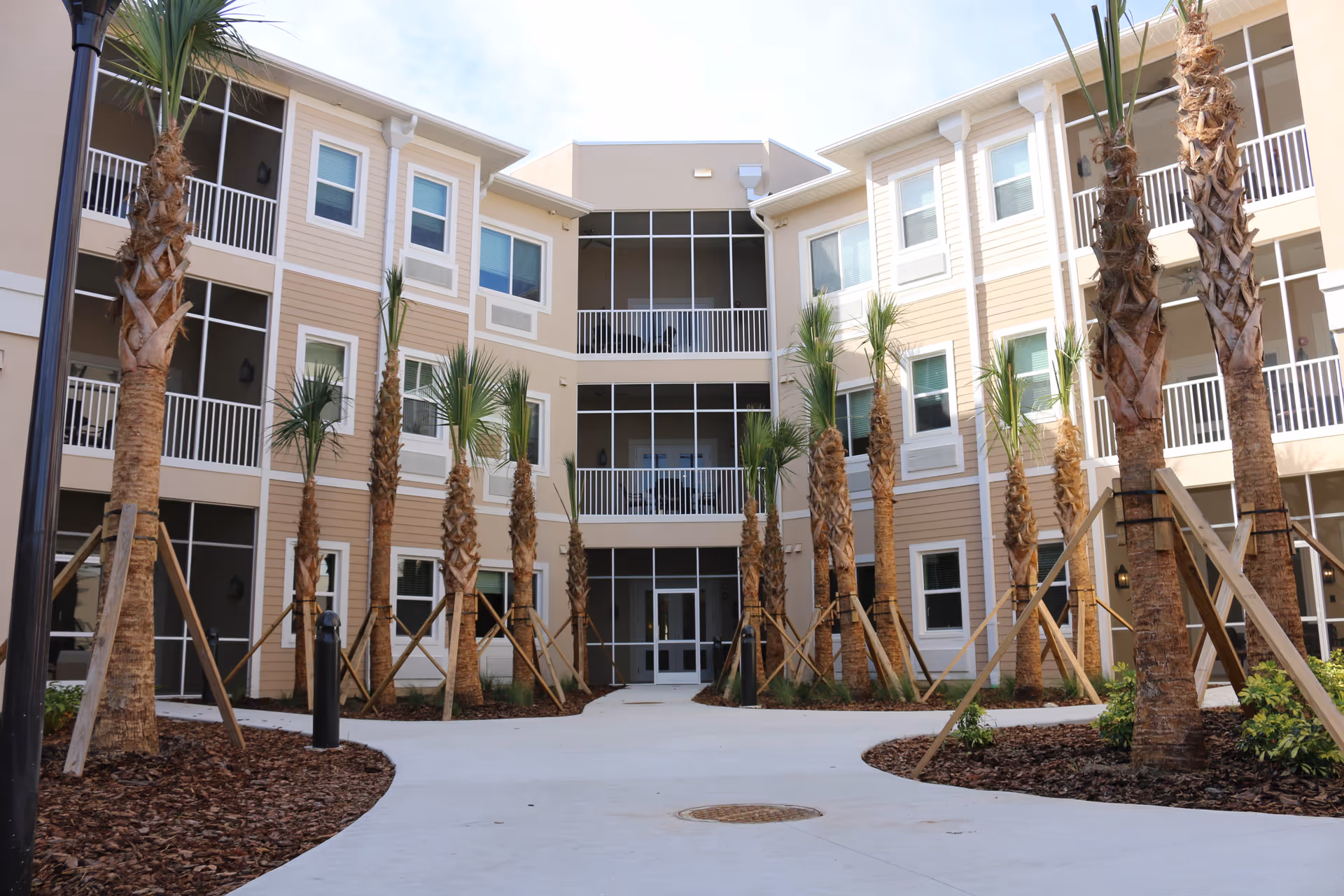 Exterior view of a three-story senior living facility building with beige and white siding, multiple windows, and balconies. The entrance is centered with glass doors, and there is a paved walkway leading to it. Young palm trees supported by wooden braces line both sides of the walkway, with mulch and small plants around their bases.