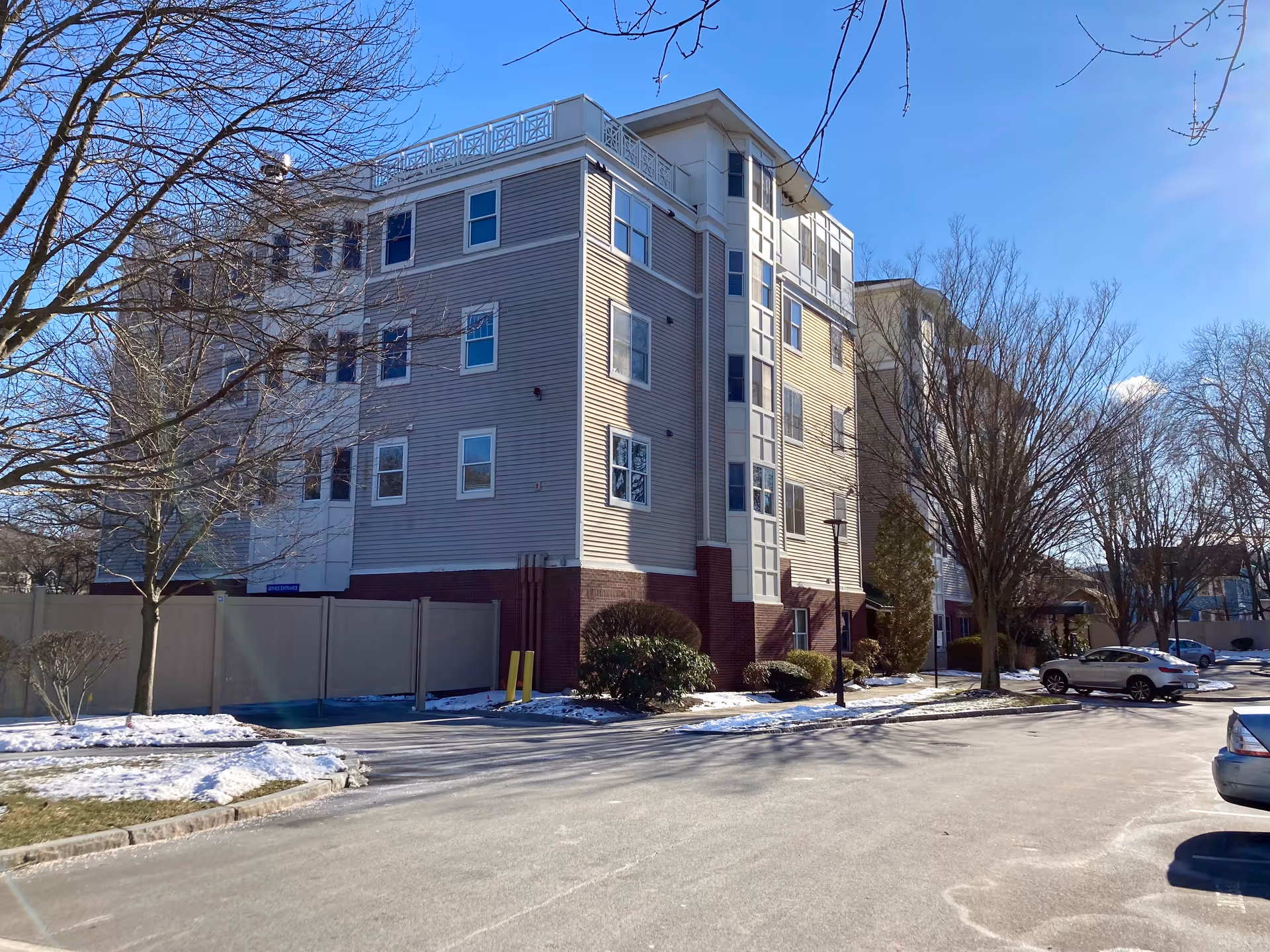 Exterior view of a multi-story senior living facility building with beige siding and red brick base, surrounded by leafless trees and patches of snow on the ground under a clear blue sky. Several parked cars are visible in the parking lot in front of the building.