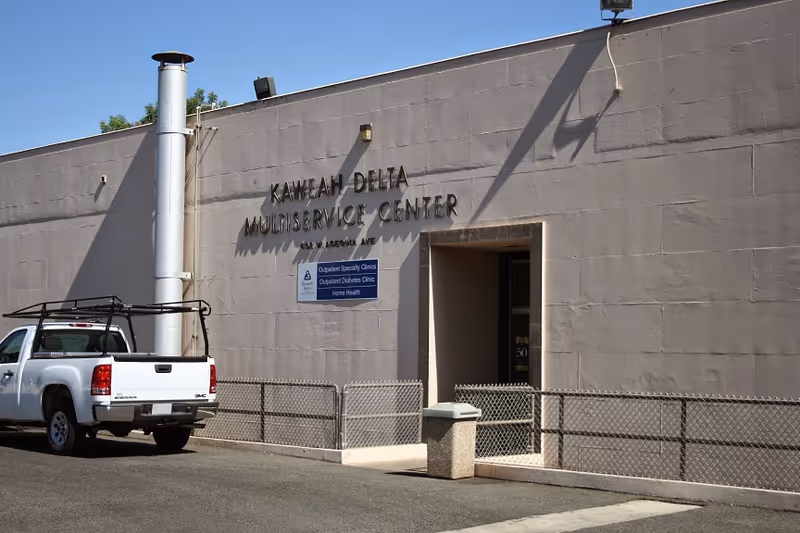 Gray building exterior with Kaweah Delta Multiservice Center signage, doorway entrance, fence, and white pickup truck.