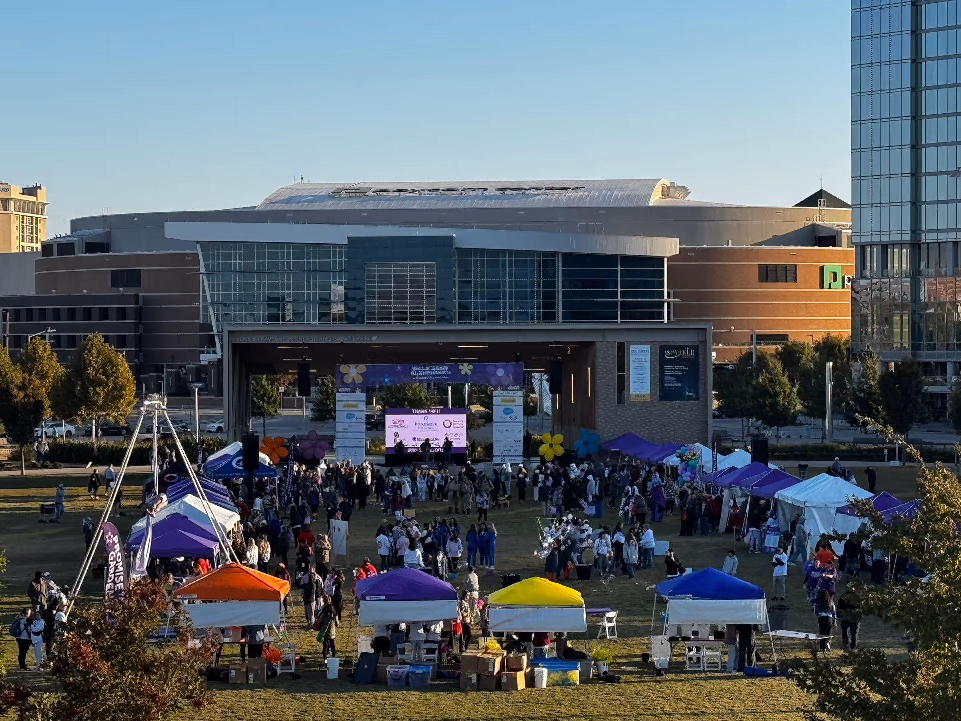 Outdoor festival with colorful tents and a stage on grassy plaza, crowds gathered.
