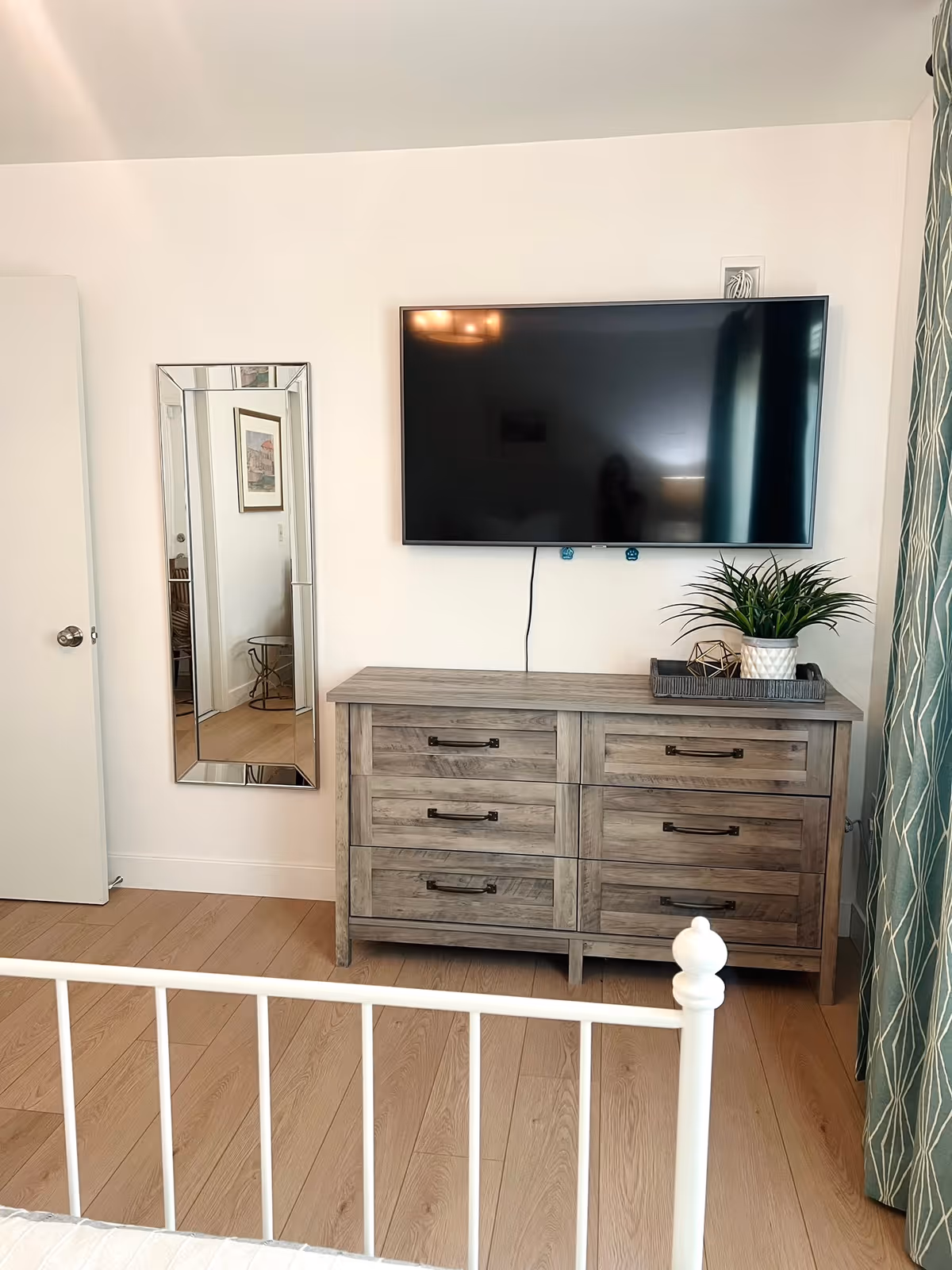 A bedroom corner featuring a wooden dresser with six drawers, a flat-screen TV mounted on the wall above it, a decorative plant in a white pot on a tray on top of the dresser, a full-length mirror mounted on the wall to the left, light wood flooring, and part of a white metal bed frame in the foreground. A green curtain with a white leaf pattern is visible on the right side.