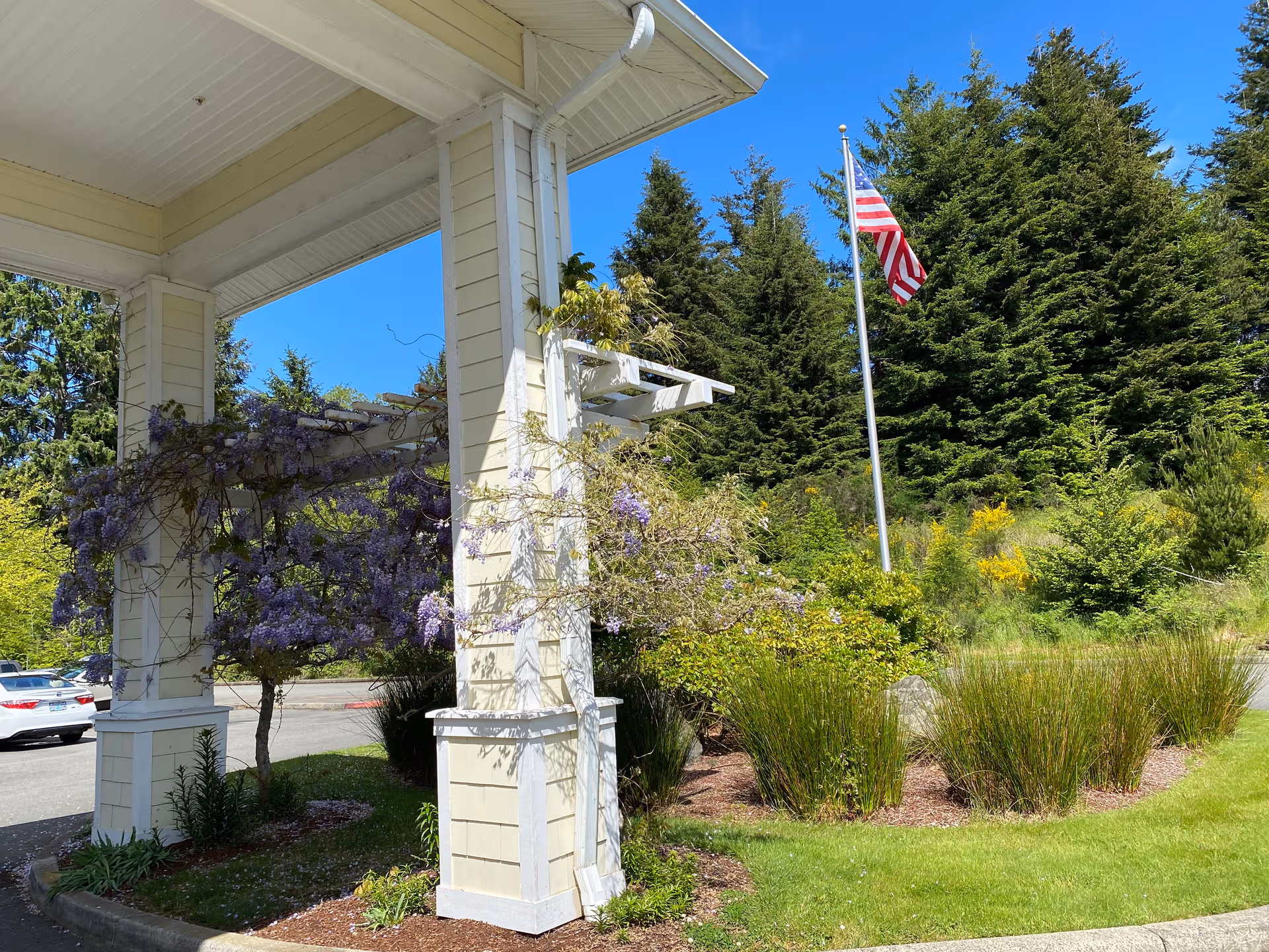 Front entrance of a senior living facility with columns, flowering vines, flag, and landscaped surroundings.