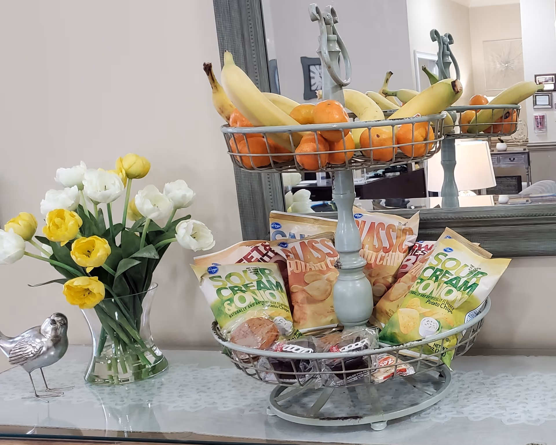 A two-tiered metal stand holding bananas and oranges on the top tier, and bags of potato chips along with some packaged snacks on the bottom tier, placed on a glass table. Next to the stand is a clear vase with yellow and white tulips and a small decorative silver bird figurine. A large mirror is in the background reflecting part of the room with a lamp and furniture.