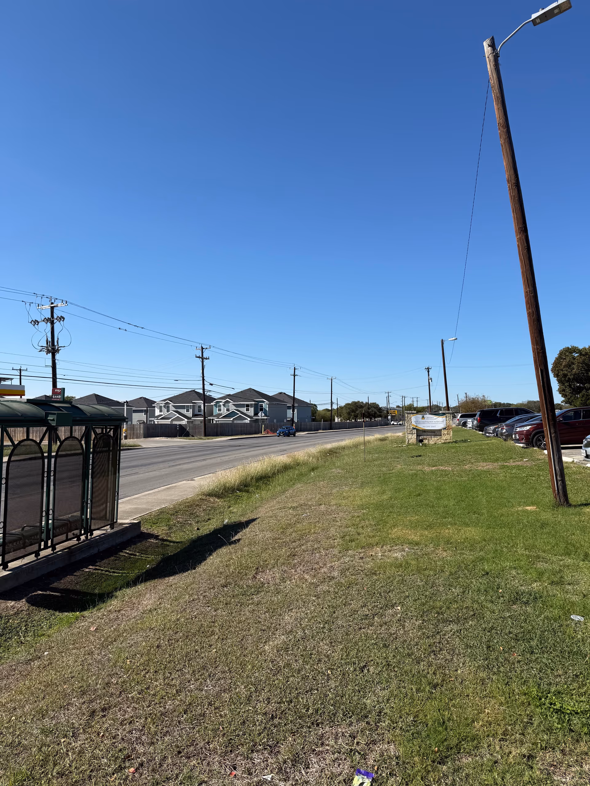 Wide outdoor scene with grassy verge, road, utility poles, and distant houses under a blue sky.