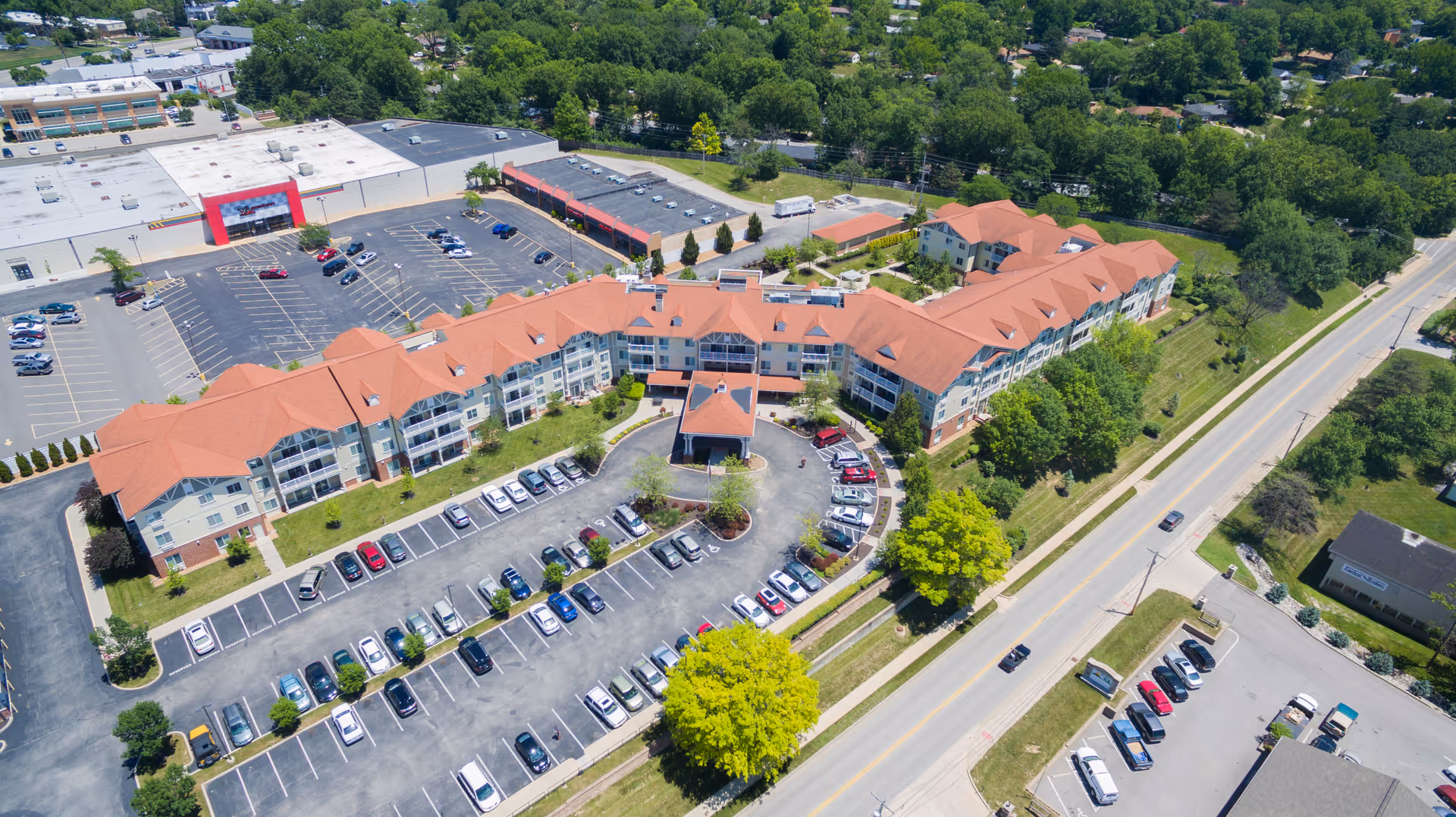 Aerial view of a large senior living facility with red roofs, surrounded by parking lots and greenery. The building is shaped in a U-formation with multiple floors and balconies. There are cars parked in the parking lots and a road running alongside the property.