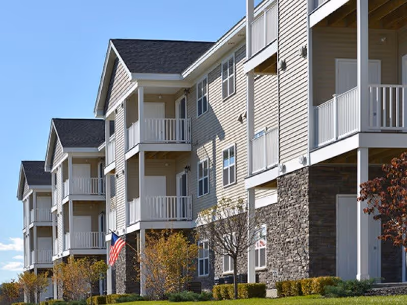 Exterior front view of a multi-story apartment building with balconies, stone accents, an American flag, and landscaped grounds.