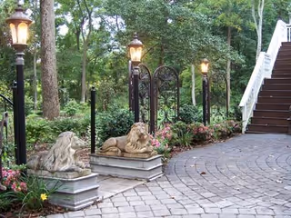 Stone statues and vintage lampposts line a curved brick path beside lush greenery.