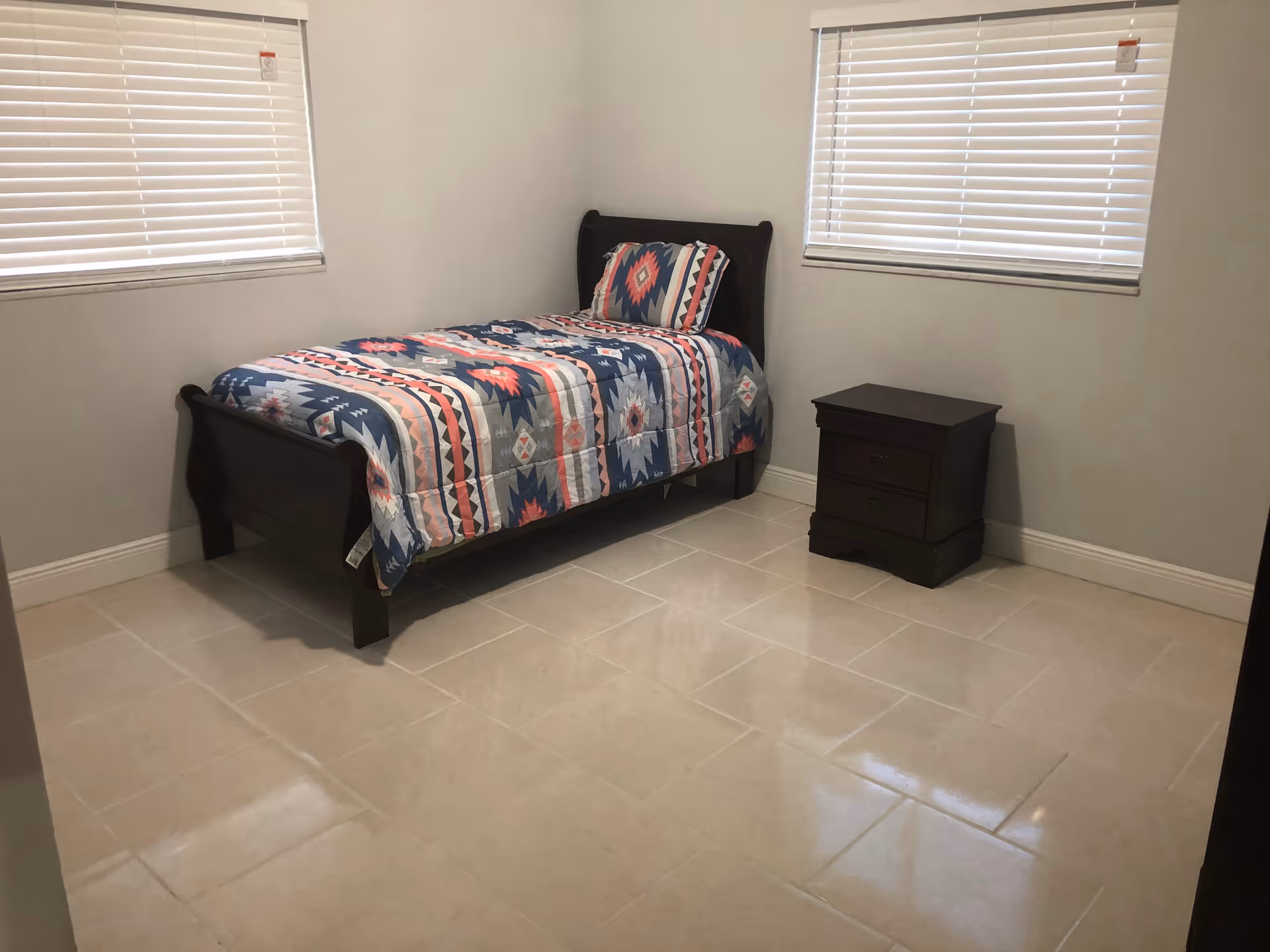 A sparsely furnished bedroom with a single bed, quilt, nightstand, and blinds.