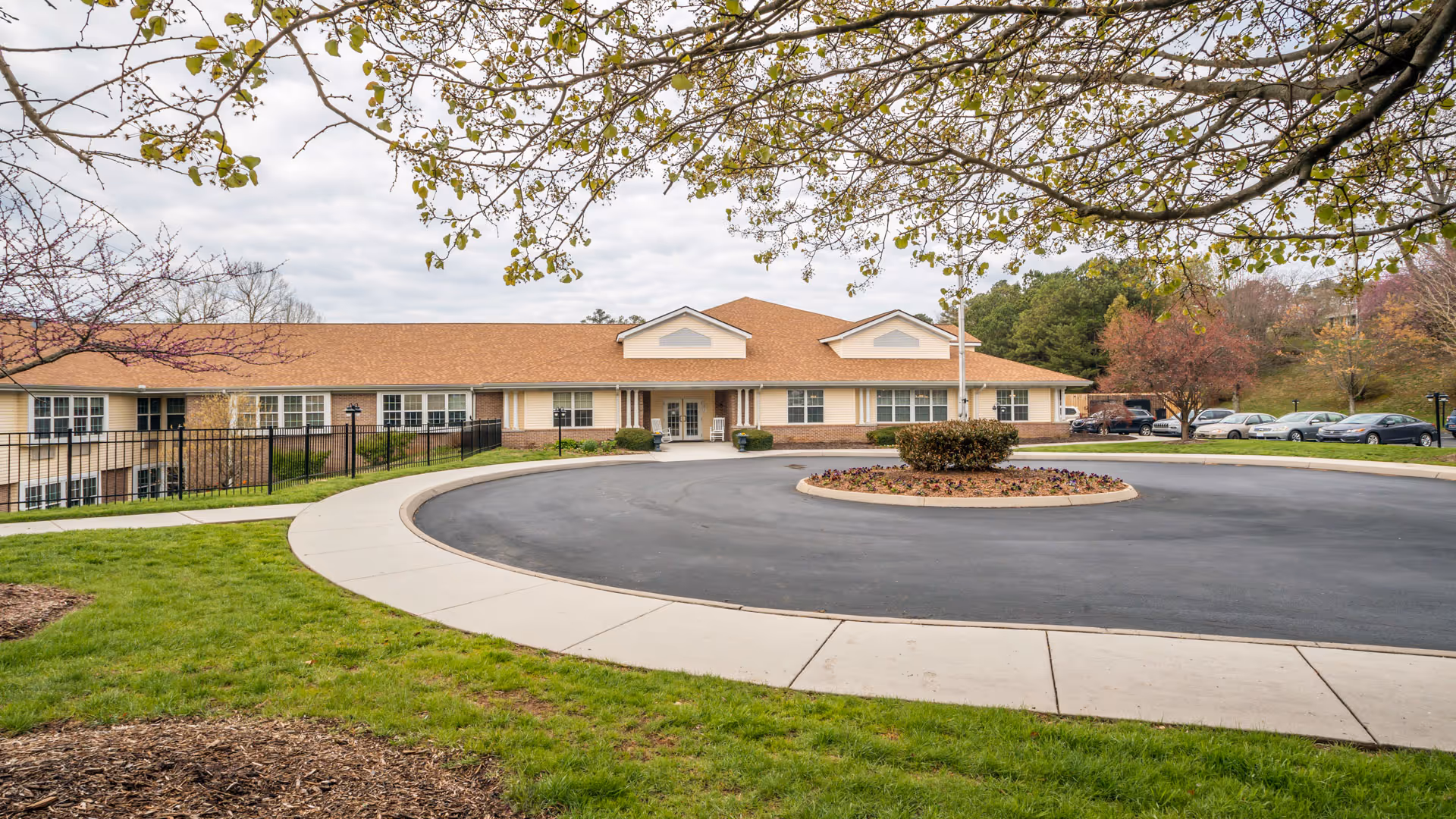 Front exterior view of a single-story senior living facility building with a circular driveway and landscaped greenery. Trees with budding leaves frame the top of the image, and several parked cars are visible to the right.