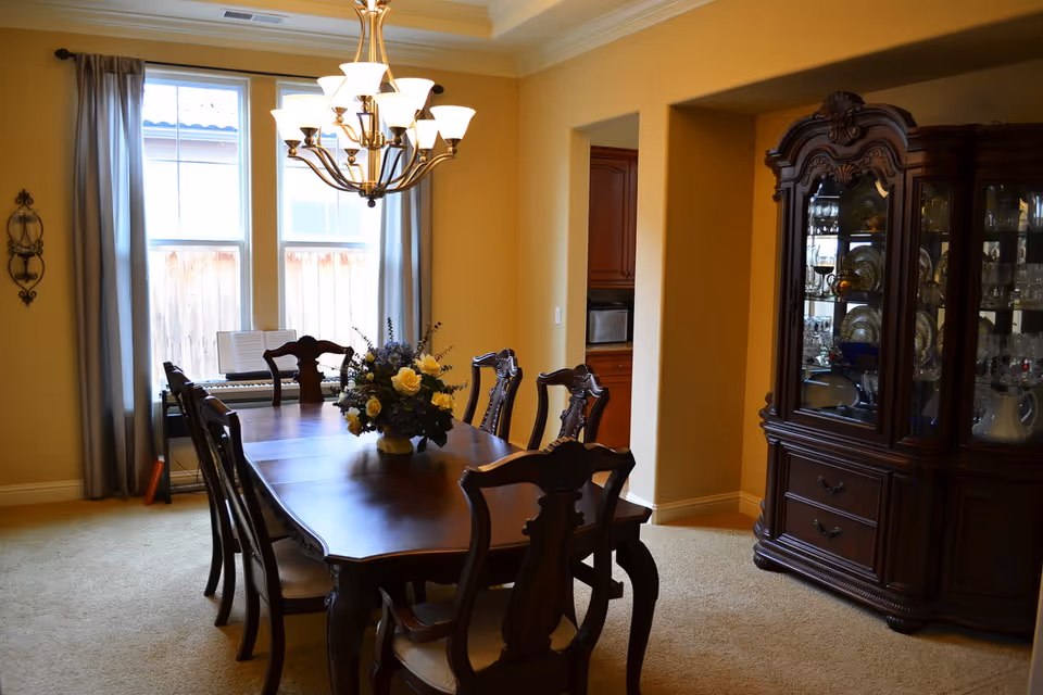 A dining room with a wooden dining table and six chairs. A floral centerpiece is on the table. There is a chandelier hanging above the table. Two large windows with gray curtains allow natural light into the room. To the right, there is a wooden china cabinet displaying glassware and dishes. The walls are painted a warm beige color, and the floor is carpeted.
