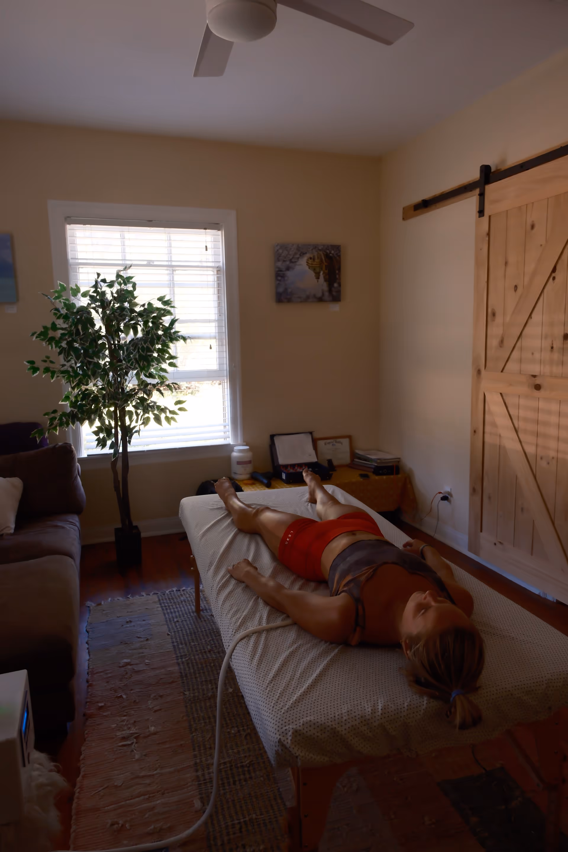 Person lies on a massage table in a room with a plant, couch, and barn door.
