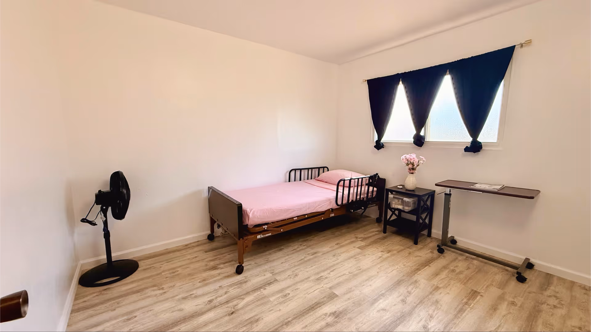 Sparse bedroom with single bed, fan, small desk, and window with dark curtains.