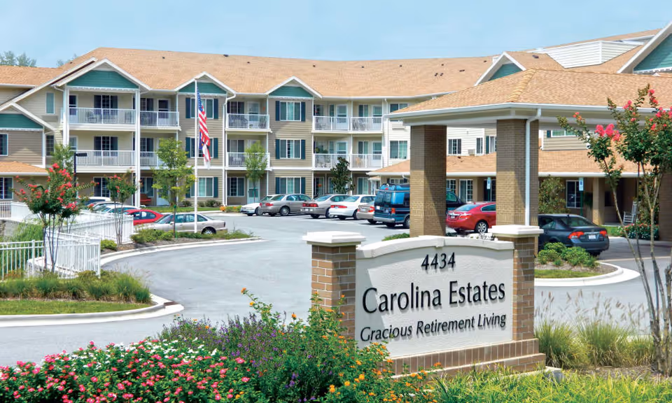 Entrance and front exterior of Carolina Estates retirement community showing the sign, landscaped grounds, parking area, and multi-story building.