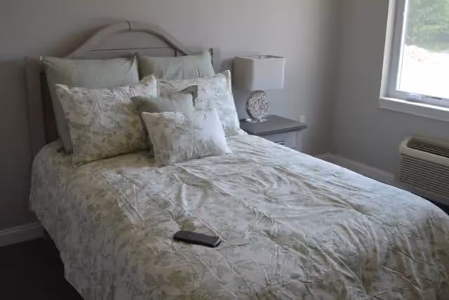 A neatly made bedroom with a floral bedspread, pillows, a nightstand, lamp, window, and air conditioner.