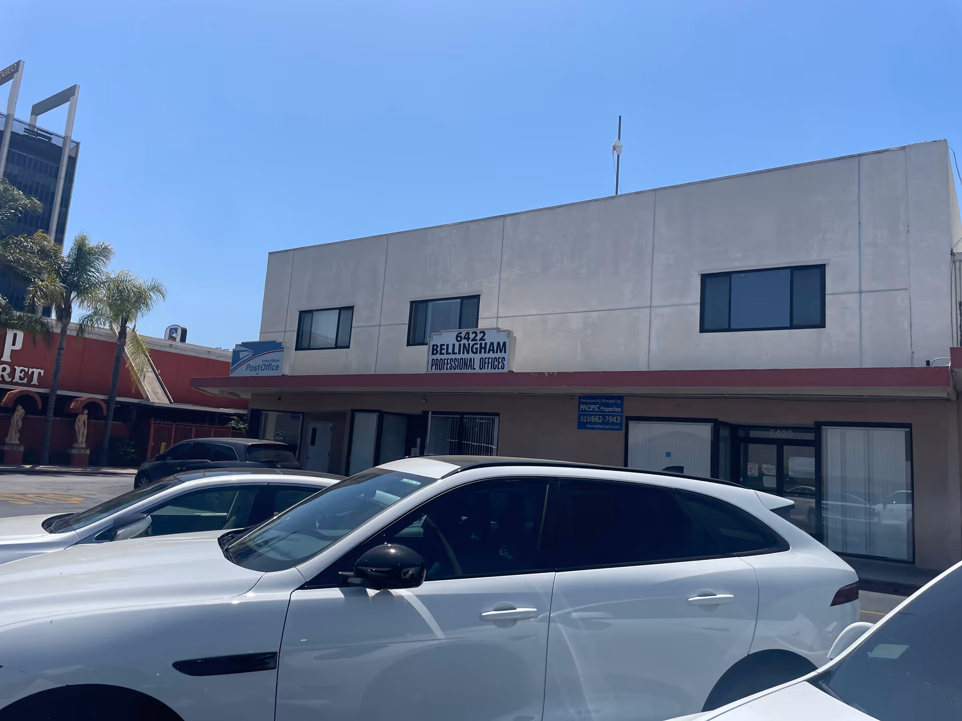 Beige, two-story office building with a sign and several parked white cars in front.