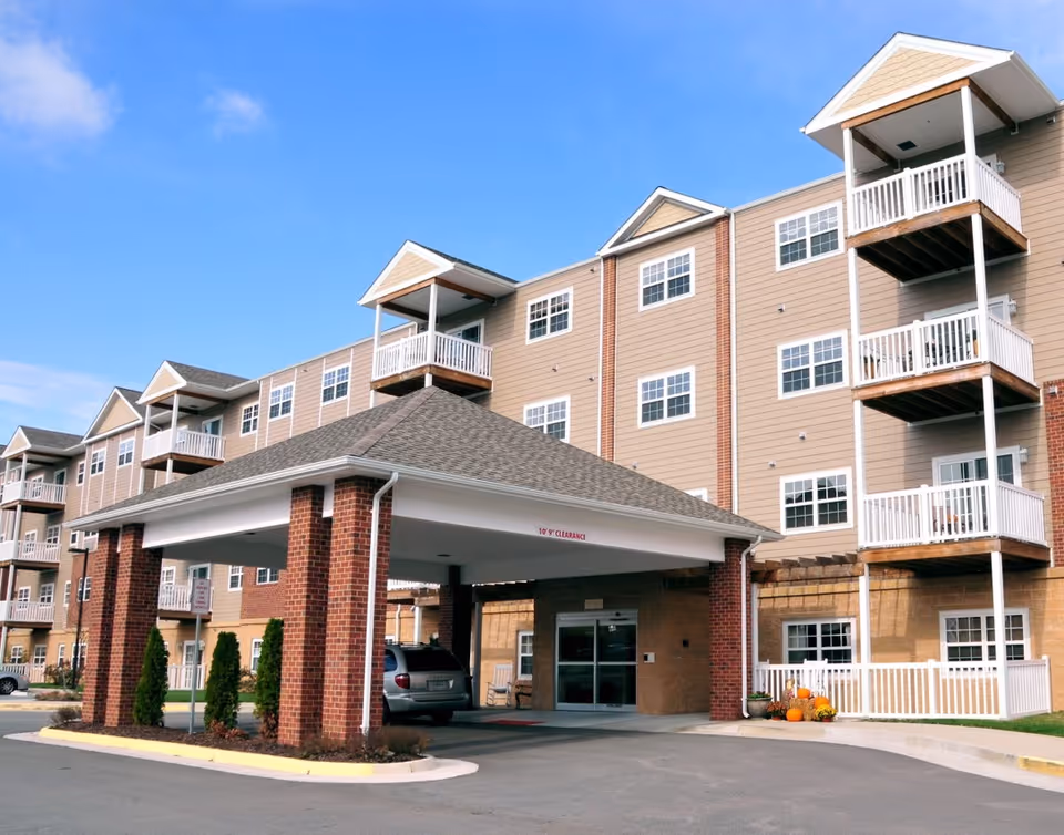 Exterior view of a multi-story senior living facility building with beige siding and brick accents. The building features multiple balconies and a covered entrance with a vehicle parked underneath. There are some small shrubs and pumpkins near the entrance, and the sky is clear with a few clouds.