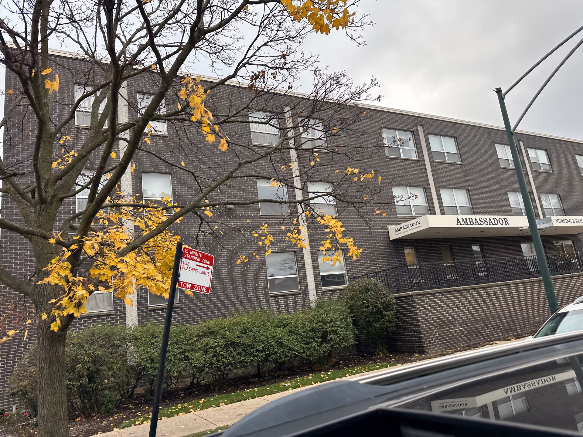 Front exterior of the three-story brick Ambassador Nursing & Rehabilitation Center with a tree, shrubs, and a street sign in the foreground.