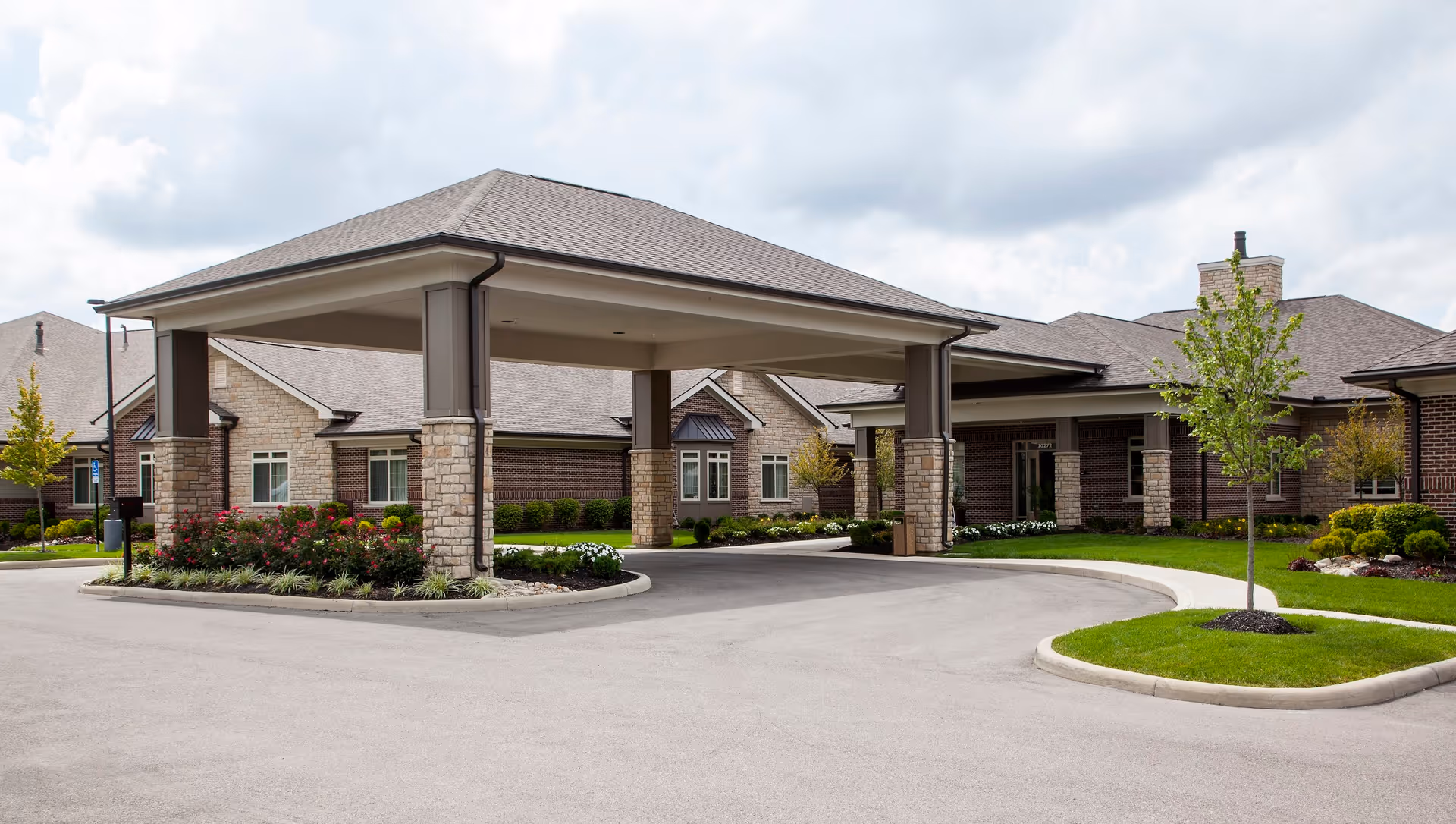 Exterior view of The Ganzhorn Suites building entrance with a covered drop-off area supported by stone pillars, surrounded by landscaped greenery and a paved driveway under a cloudy sky.