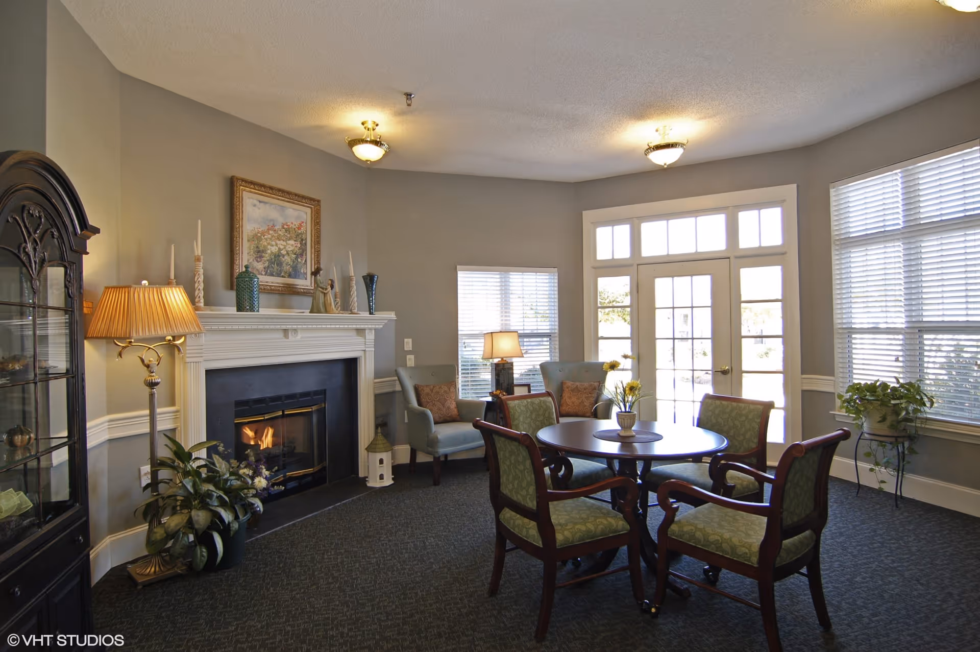 Sunlit common room with a round table and four chairs, armchairs by a fireplace, and large windows and French doors.