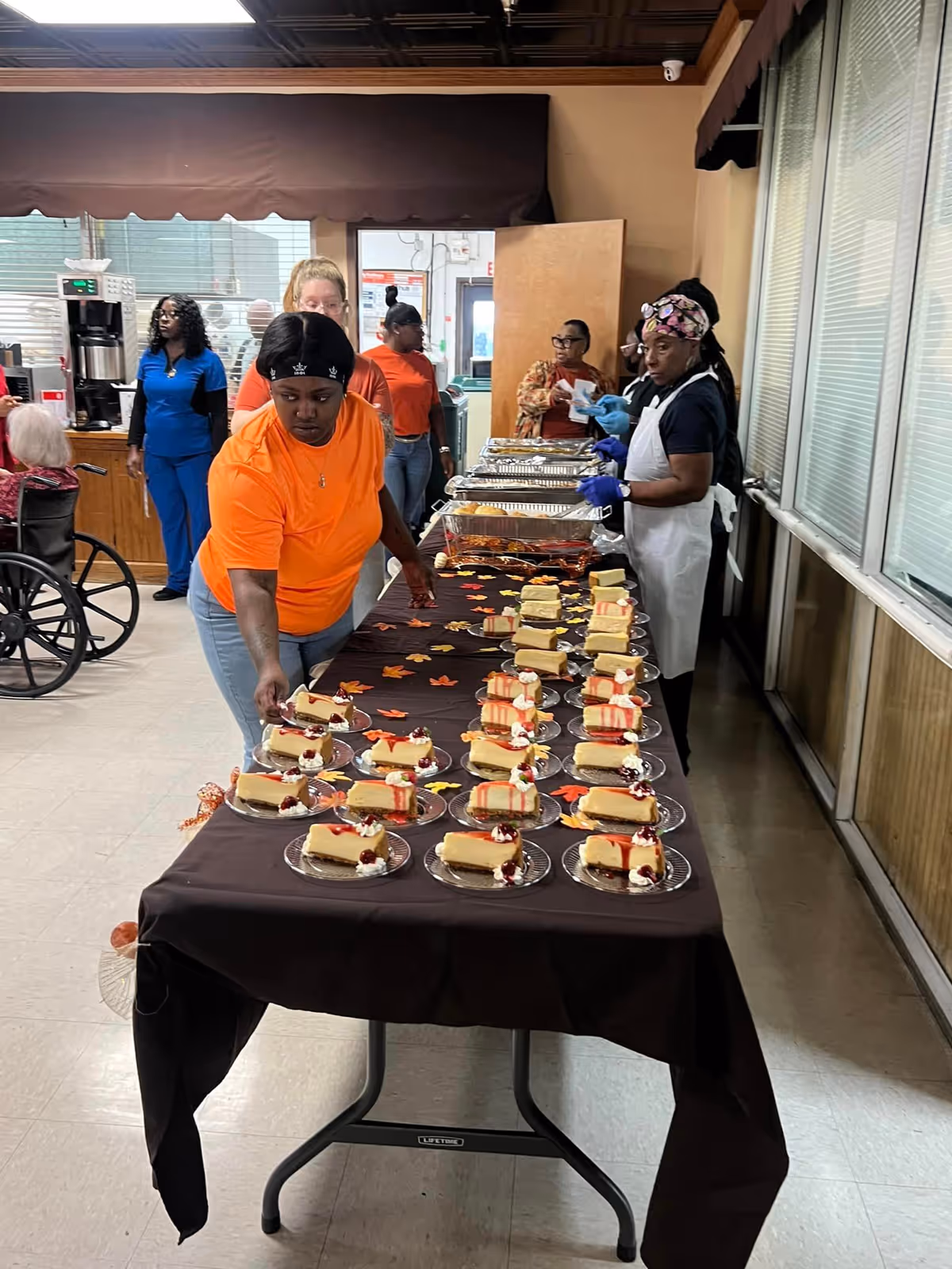 Several staff and residents set out plated slices of cake along a long table in a dining area.