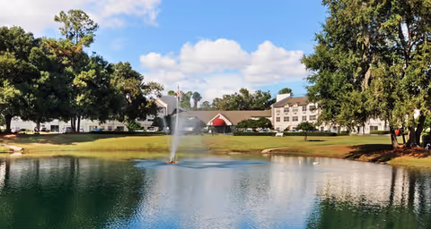 A large pond with a water fountain in the center is surrounded by green grass and trees. In the background, there is a multi-story building with a red entrance canopy, likely part of a senior living facility. The sky is blue with scattered clouds.