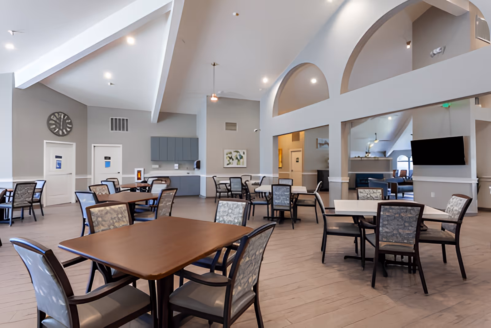 Spacious dining area in an assisted living facility with multiple tables and chairs arranged neatly. The room features high vaulted ceilings with exposed beams, a large wall clock, and a mounted flat-screen TV. There are cabinets and a countertop along one wall, and arched openings lead to an adjacent lounge area with sofas.
