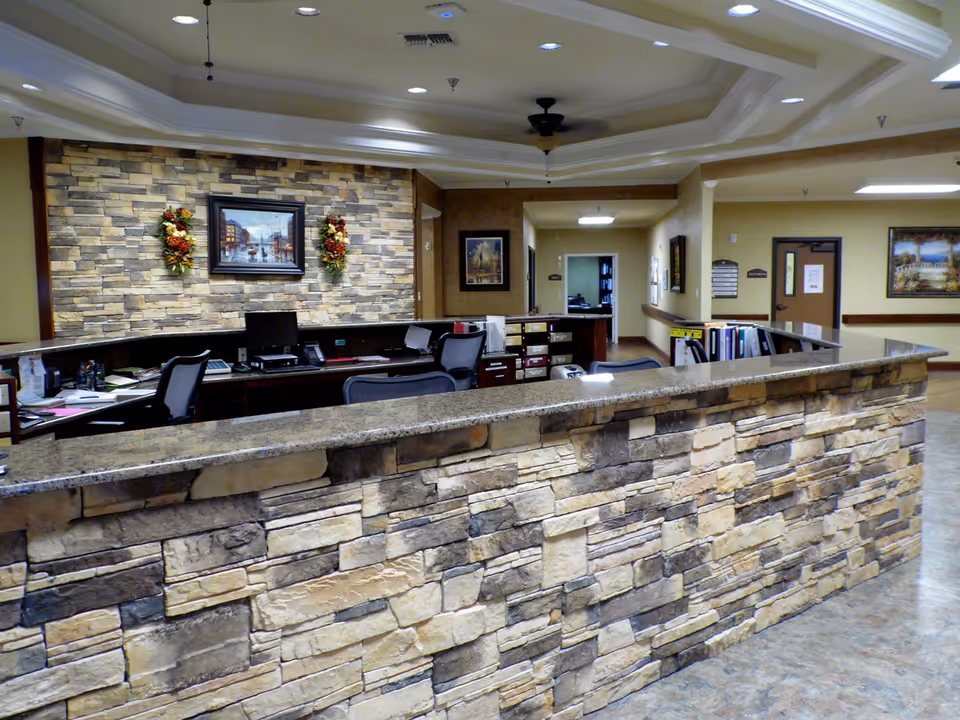 Reception area with a stone facade counter and granite top, office chairs, computer monitors, and decorative wall art including a framed painting and floral arrangements on a stone accent wall.
