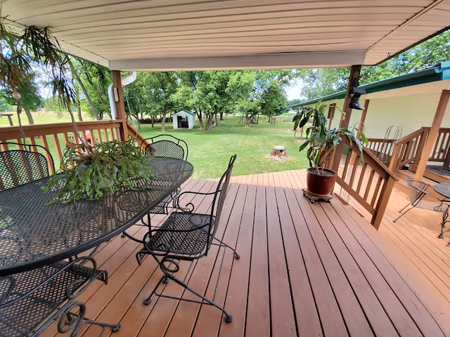 Covered wooden deck with a metal patio table and chairs overlooking a grassy yard with trees and a small shed.