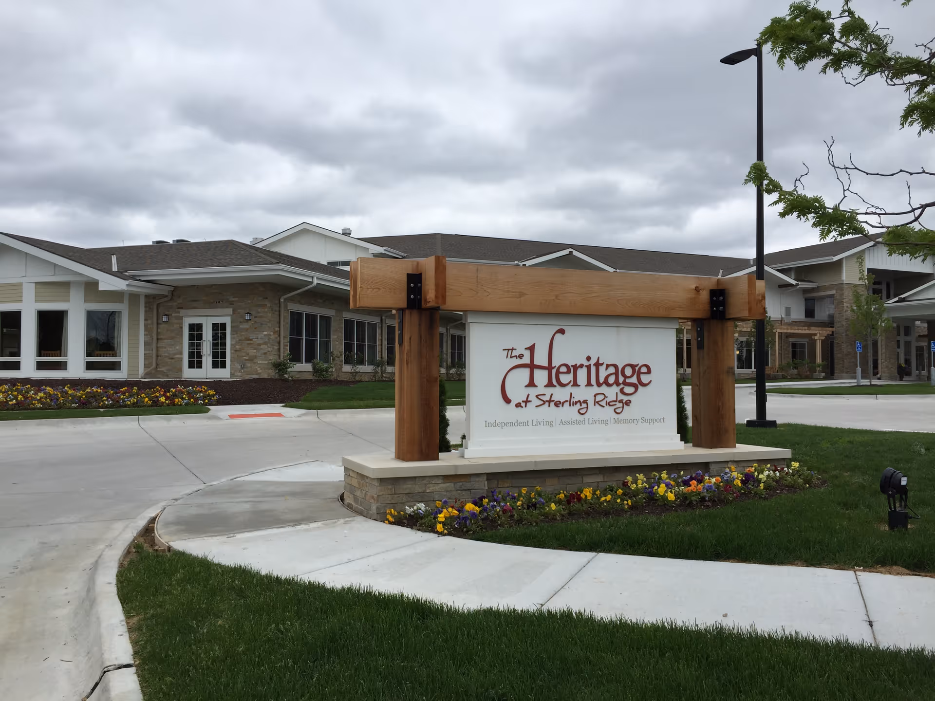 Exterior view of The Heritage at Sterling Ridge senior living facility with a large sign in front displaying the facility's name and services. The building has a modern design with stone and white siding, surrounded by a well-maintained lawn and flower beds under a cloudy sky.