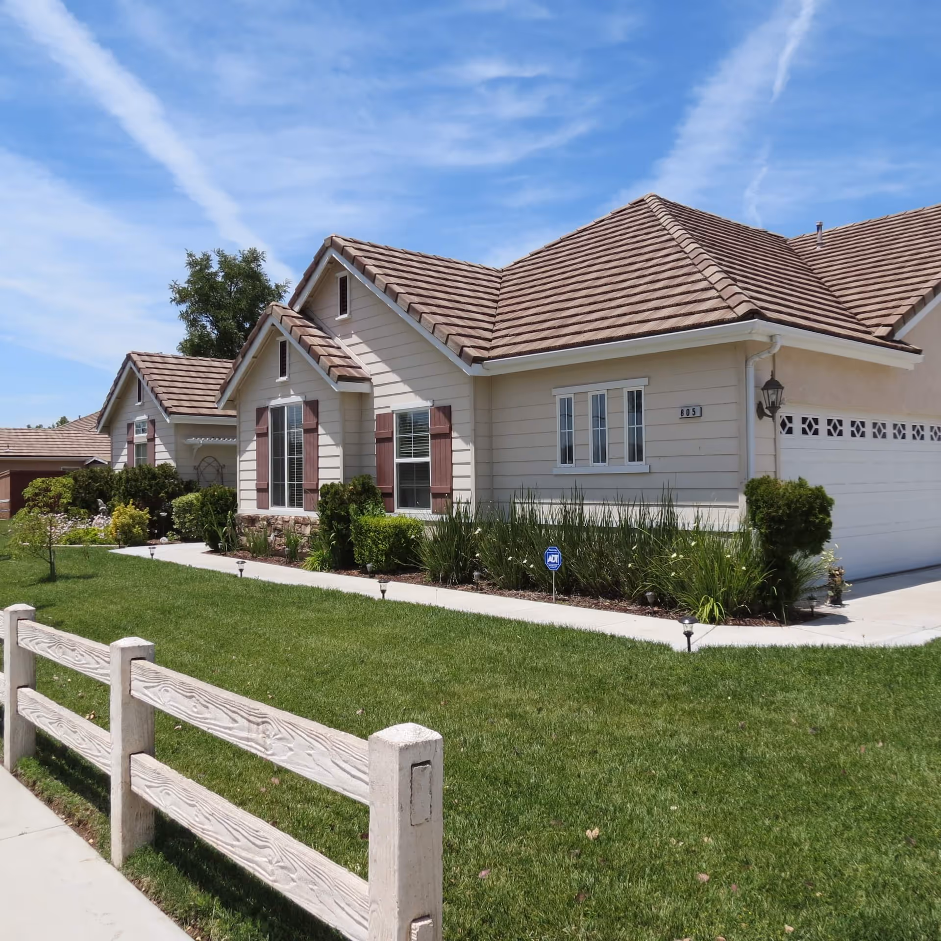 Exterior view of a single-story residential building with beige siding, brown tiled roof, and white trim. The house has multiple windows with brown shutters, a well-maintained green lawn, shrubs, and a white wooden fence along the sidewalk under a blue sky with wispy clouds.