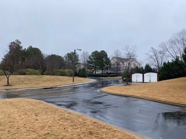 Wet paved road curving through a landscaped area with dry grass and scattered trees under an overcast sky, with a large building partially visible in the background.