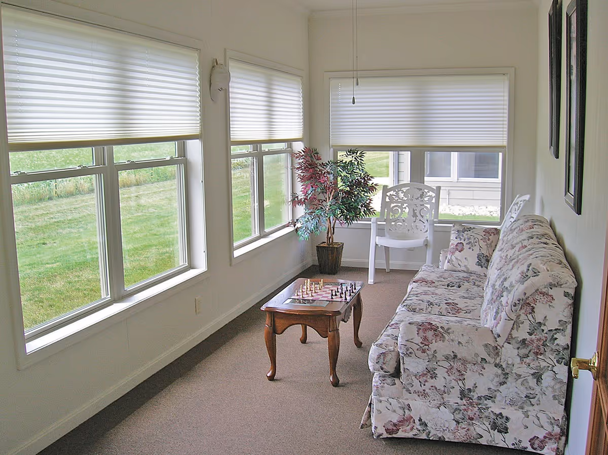 Bright sunroom with large windows, floral sofa, chessboard on a small coffee table, and white chairs.