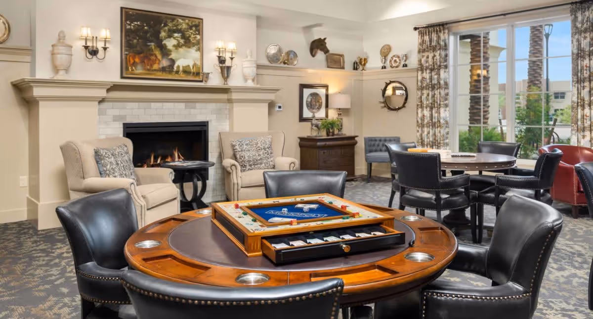 A cozy senior living common area with a round wooden game table featuring a board game, surrounded by black leather chairs. In the background, there are two beige armchairs with patterned pillows near a lit fireplace, decorative wall sconces, and various wall decorations including a painting and a mounted animal head. Large windows with floral curtains let in natural light and show palm trees outside.