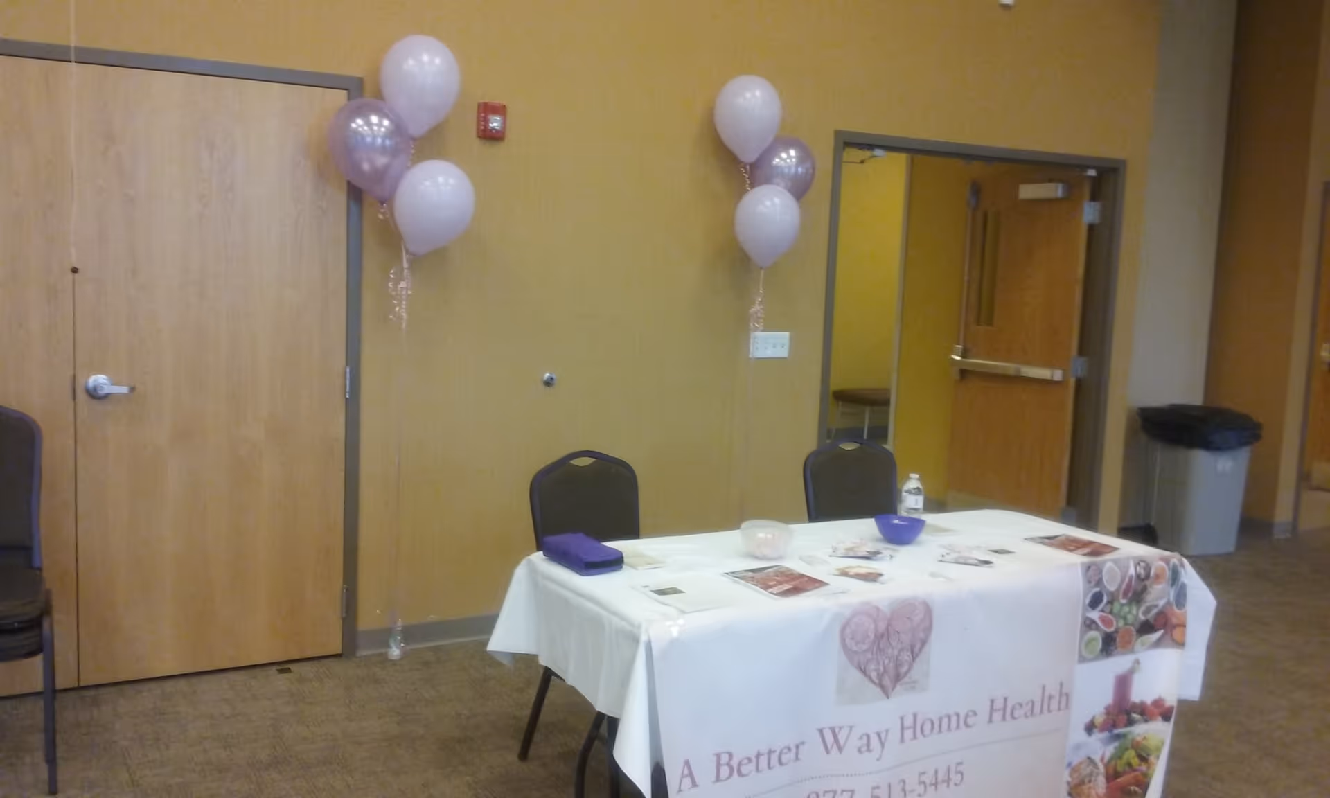Indoor event setup with a bannered table, balloons, and empty chairs in a beige room.