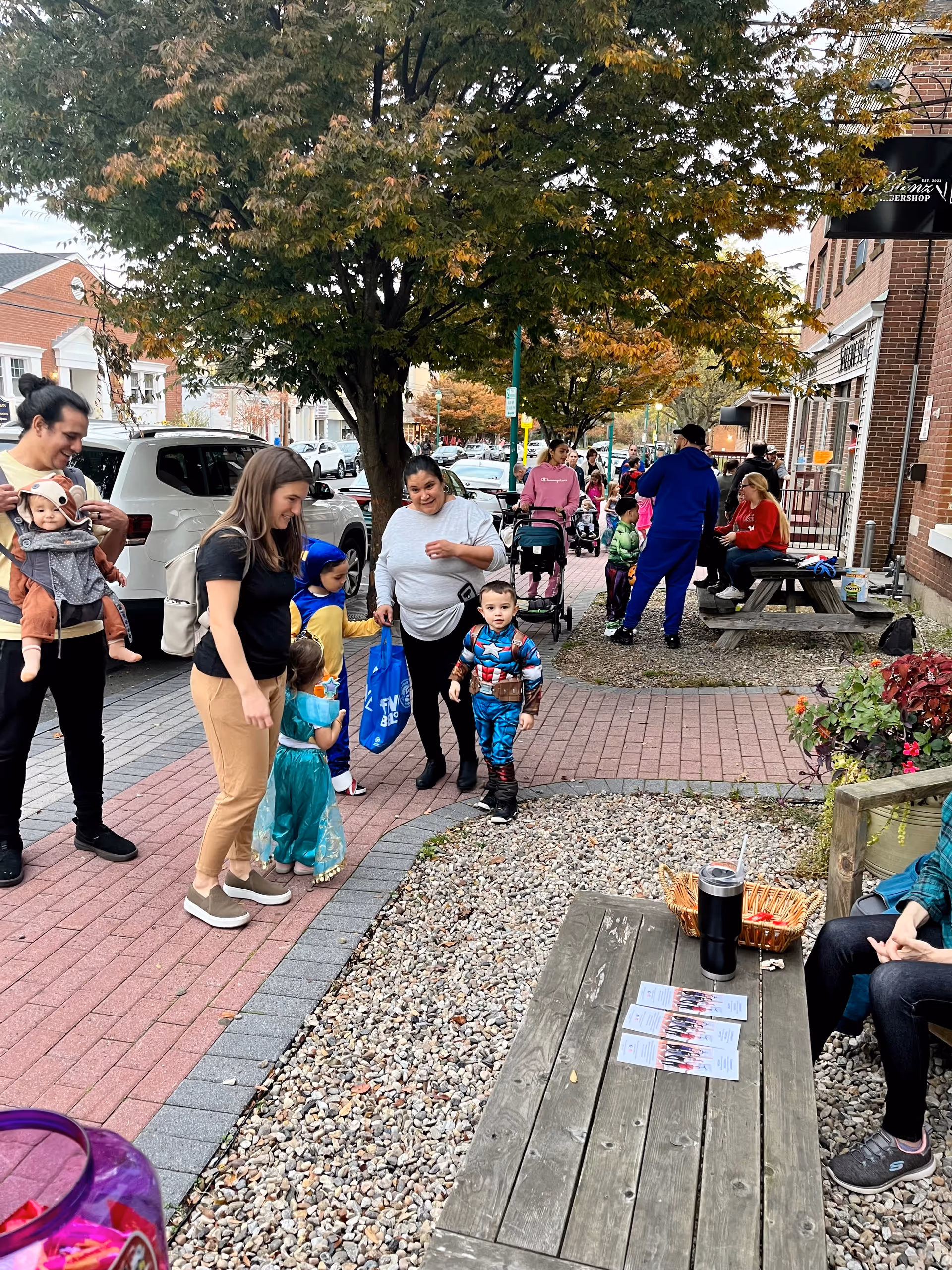 Outdoor street scene with families and children gathered near a brick building and picnic table.