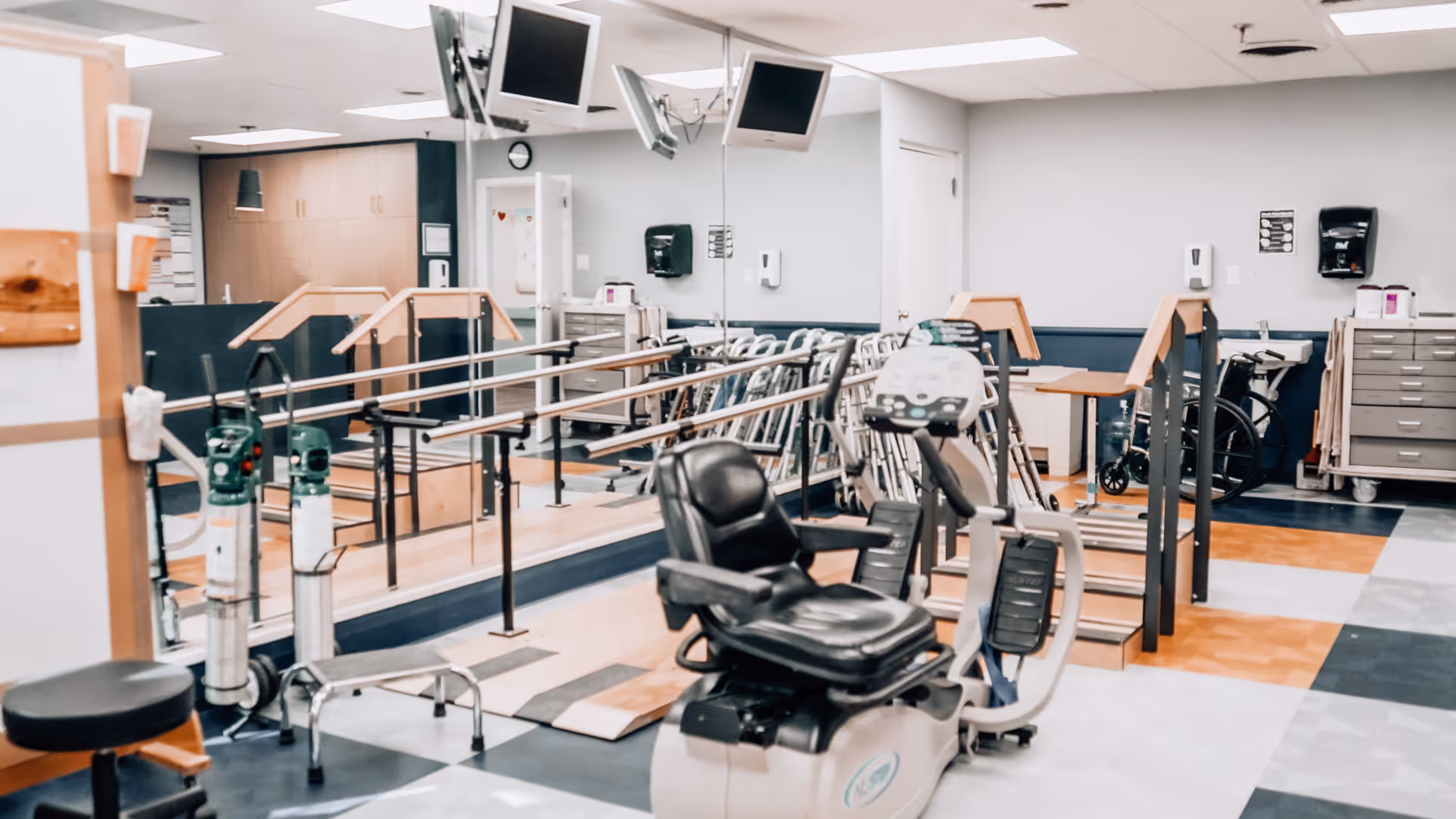 Physical therapy room with exercise equipment including a recumbent bike, parallel bars for walking practice, walkers, wheelchairs, and oxygen tanks. The room has a large mirror on one wall and a tiled floor with a mix of colors.
