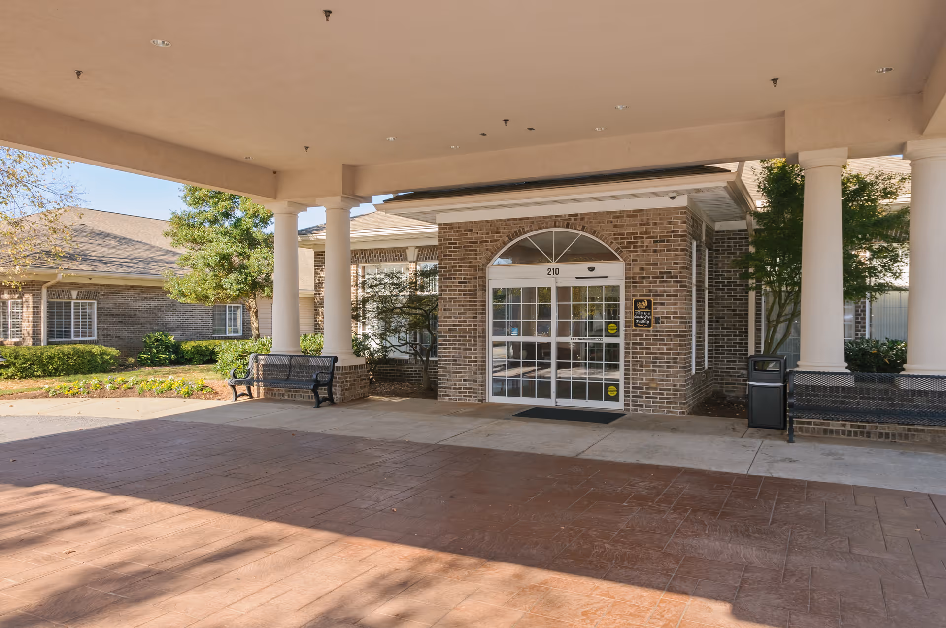 Covered entrance area of a brick building with white columns, benches, and glass double doors labeled 210. There are trees and shrubs around the building under a clear sky.