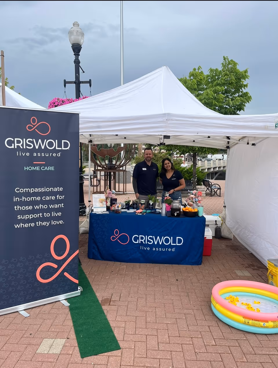Two Griswold Home Care representatives stand at an outdoor information booth under a white canopy.