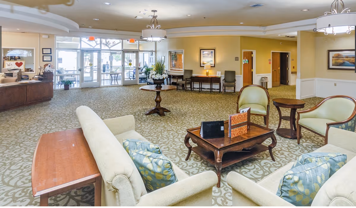 Spacious, well-lit senior living facility lobby with seating area, coffee table, reception desk, and glass entry doors.