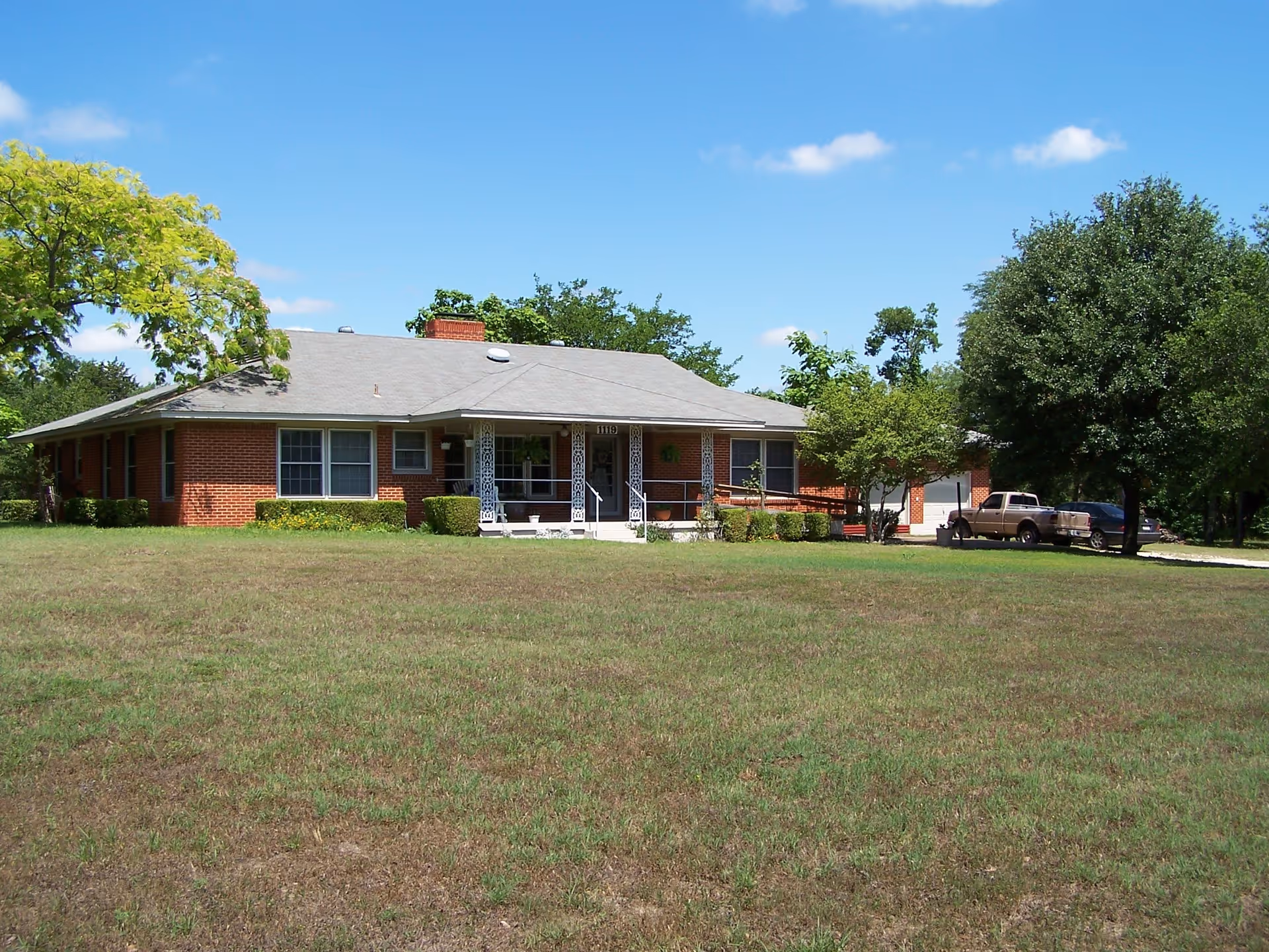 Single-story brick ranch-style house with a wide front lawn, covered porch, and trees under a blue sky.