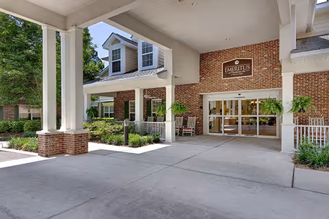 Covered entryway and portico of a brick senior-living building with white columns, hanging ferns, rocking chairs, and sliding glass doors.