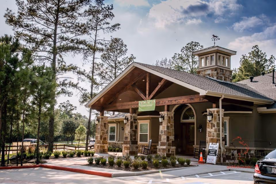 Exterior view of a single-story building with stone pillars and a wooden roof overhang at the entrance, surrounded by trees and landscaping. There is a sign near the entrance indicating valet parking and a small parking area with a car visible on the right side.