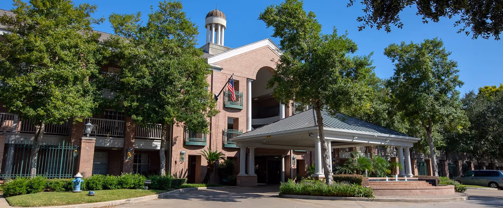 Front entrance of a brick senior living building with a covered porte-cochère, trees, and an American flag.