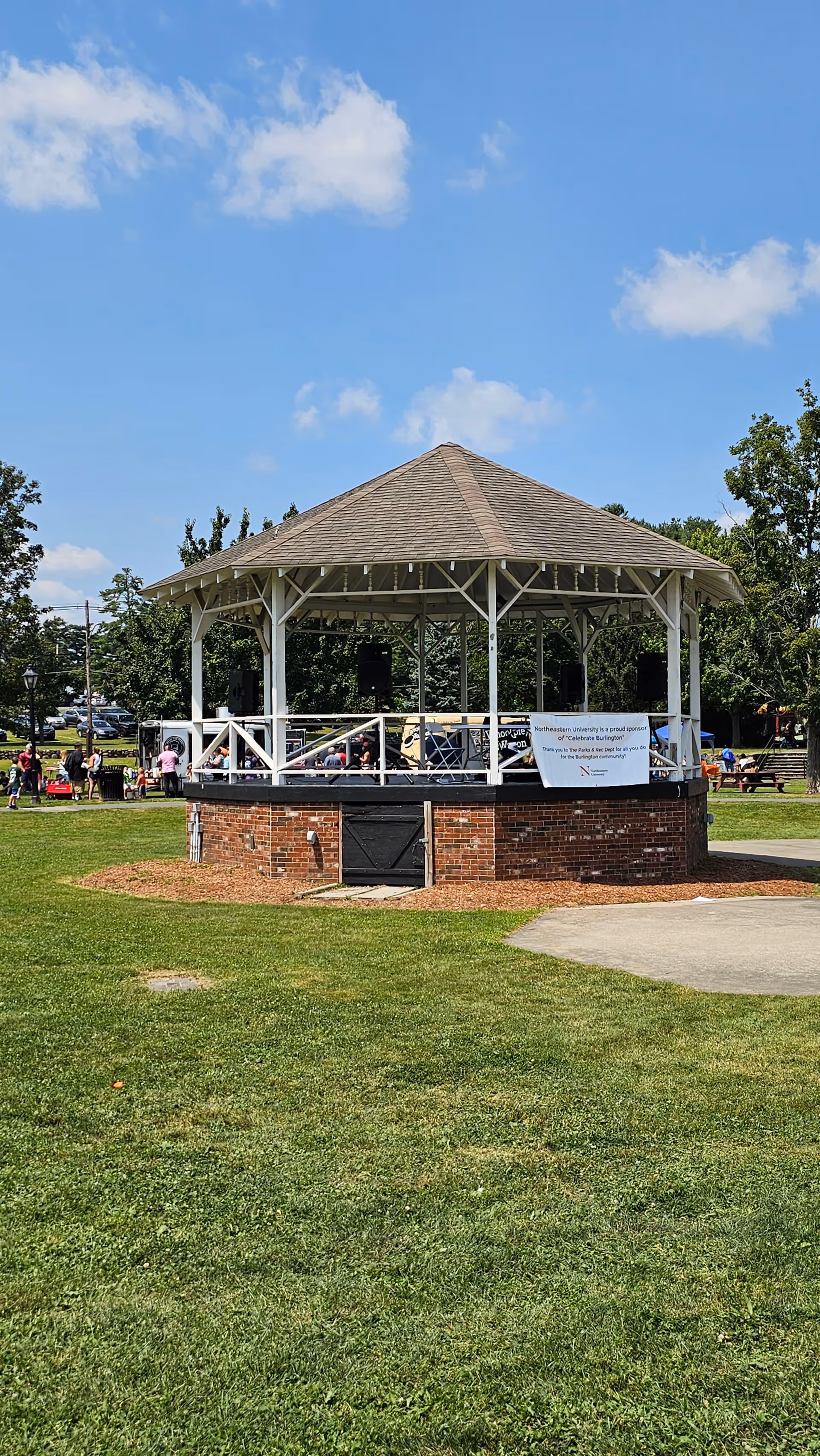 A white gazebo with a brick base sits on a grassy park area under blue sky.