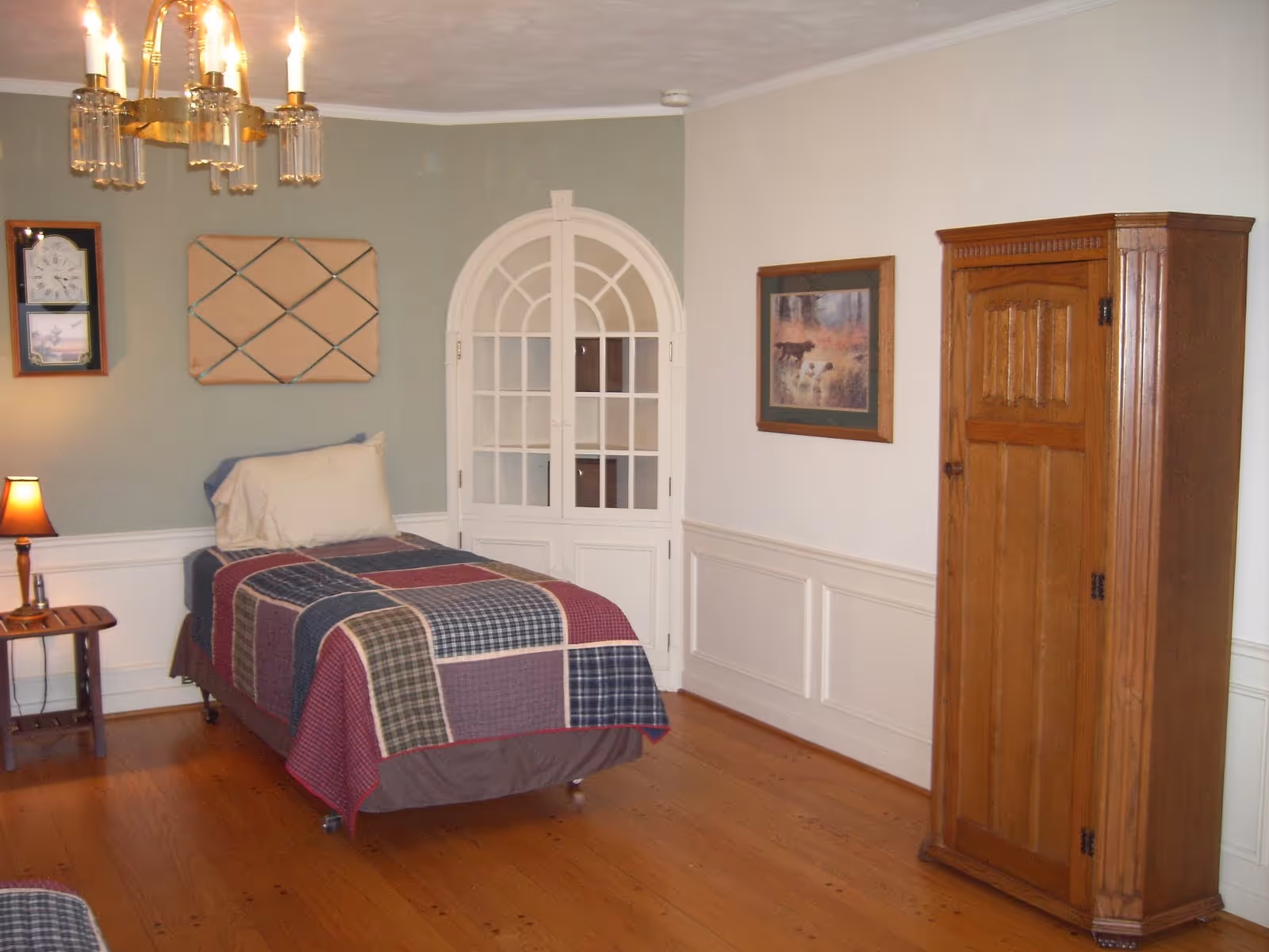 A simple bedroom with a single bed, quilt, wooden wardrobe, arched window door, and lamp.