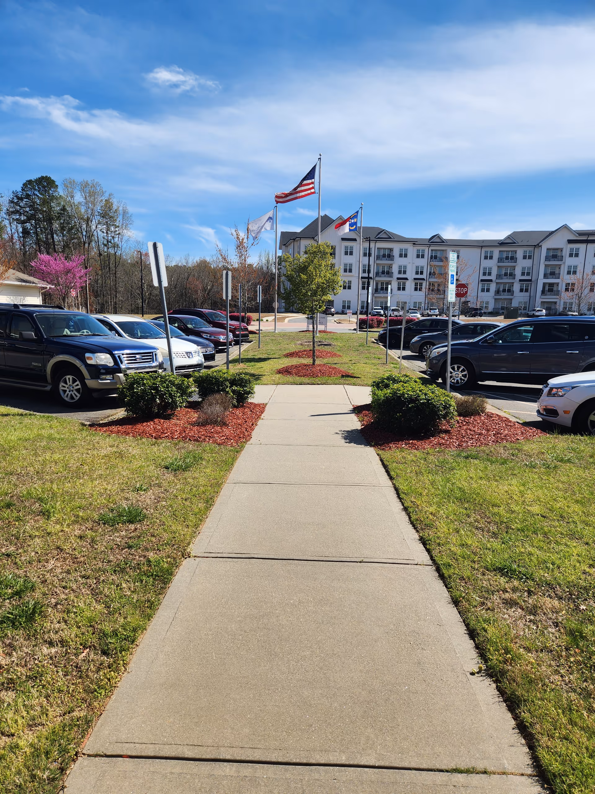 Wide walkway leads to a multi-story senior living building with flags and cars.