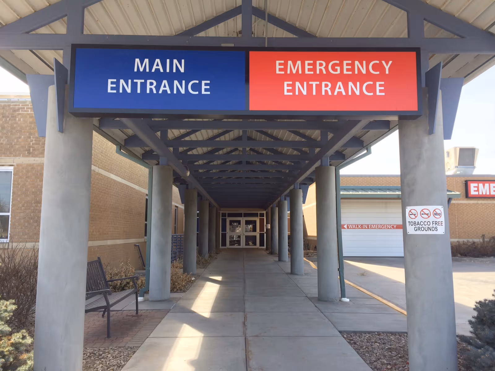 Exterior hospital entrance with blue main and red emergency signs under a covered walkway.