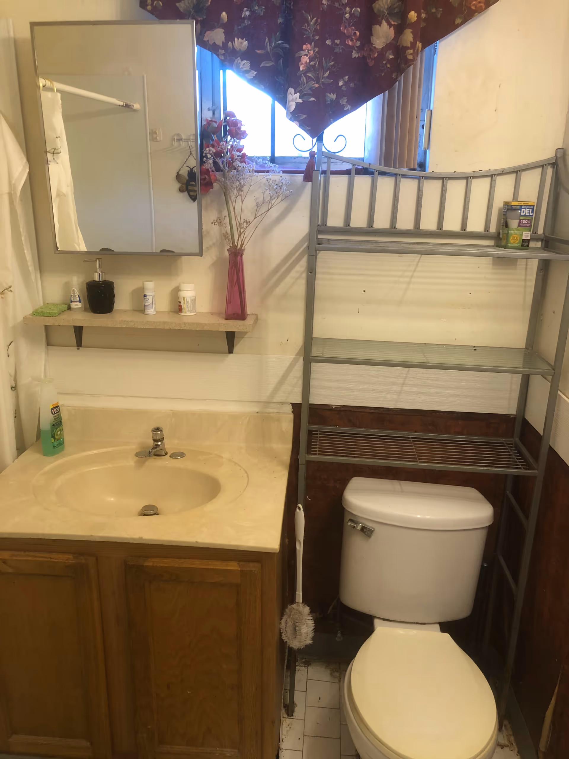 Small bathroom with sink, mirror, toilet, metal shelving, and a window with floral curtain.