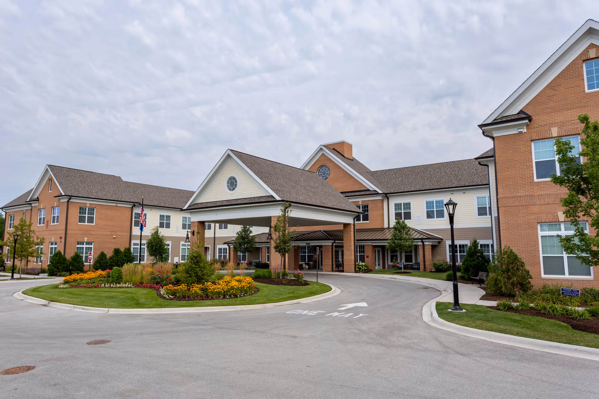 Exterior view of Arbor Terrace Glenview senior living facility showing a large brick and siding building with a covered entrance, landscaped flower beds, trees, and a driveway with a one-way arrow.